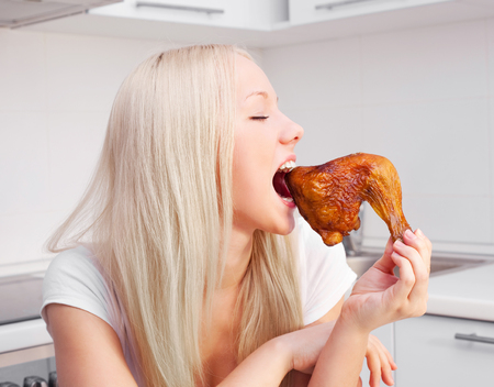 beautiful young blond woman eating fried chicken at home in the kitchenの写真素材