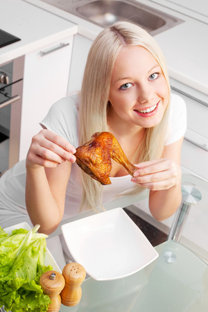 beautiful young blond woman eating fried chicken at home in the kitchenの写真素材