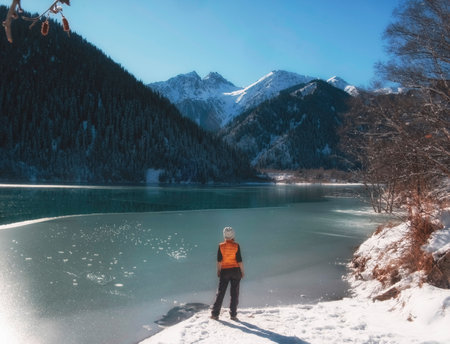 A woman tourist admires a beautiful winter lake in the mountains, surrounded by a spruce forest with white fluffy snow. Lake Issyk Almaty Kazakhstan.の写真素材