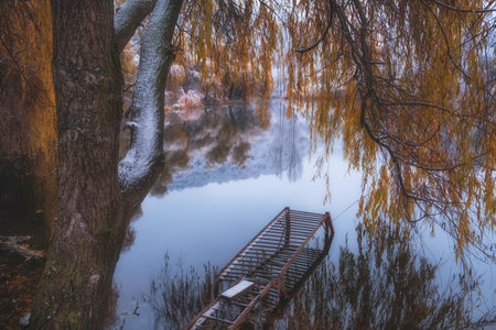 A fishing pier on a lake in late autumn, when the yellow leaves have not yet fallen from the trees, but the snow has already fallen and there is a reflection on the waterの写真素材