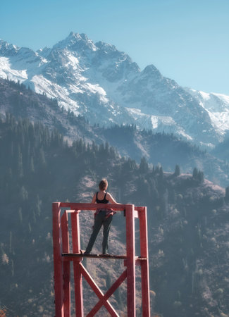 a woman standing on an observation deck, admiring the stunning view of snow capped mountains and forest on a sunny day.の写真素材