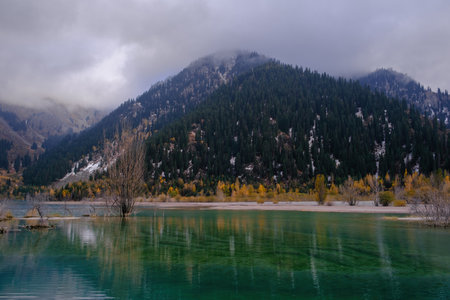 Lake Issyk in autumn. The calm surface of the water reflects the slopes of the mountains, covered with coniferous forest and yellowed trees.の写真素材