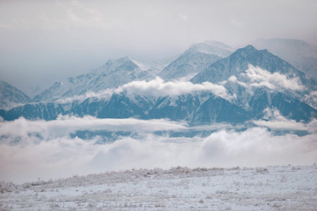 The snow-capped peaks of the Trans-Ili Alatau in the Almaty region, surrounded by thick white clouds. Scenic view of the highlands in winter: frozen steppe in the foreground and powerful mountain ranges. The atmosphere of grandeur, tranquility and pristine nature of Kazakhstan. Ideal for hiking and winter backgroundsの写真素材