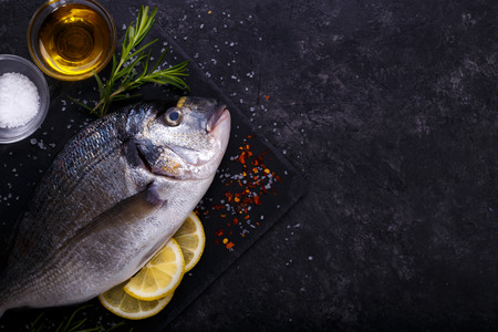 Fresh raw sea bream fish with lemon slices, salt, rosemary and olive oil on black slate background. Healthy food concept, top view, copy spaceの写真素材