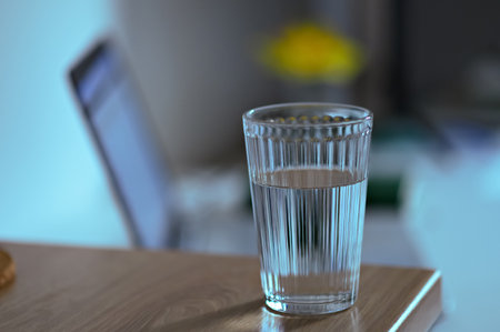 A glass filled with clear water sits on a wooden surface next to a blurred laptop, creating a refreshing and tranquil atmosphere.の写真素材