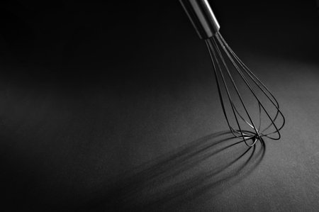 A metal whisk lies on a matte black surface, creating delicate shadows that enhance the simplicity...