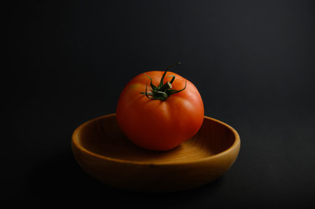 A ripe tomato with vibrant red color sits in a smooth wooden bowl, illuminated against a dark...