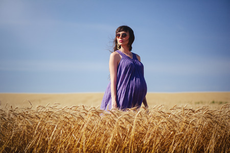 beautiful pregnant woman in a wheat field glasses and dress looking into the distance on a blue sky backgroundの写真素材