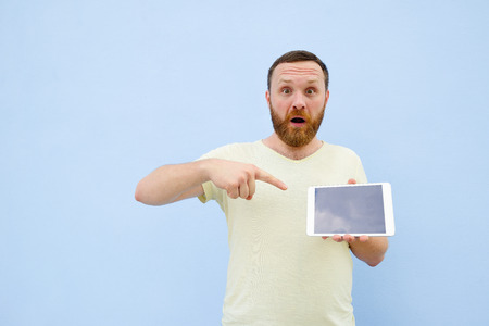 Upset Handsome young man with a beard showing something on a tablet isolated on a blue background, bright and light.の写真素材