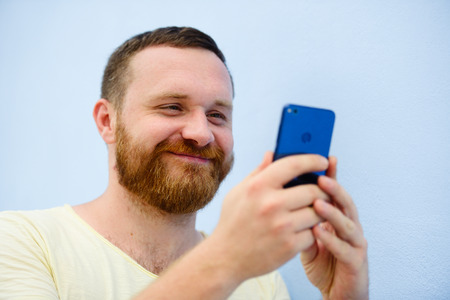 cheerful man with a red beard looks into the phone on a blue background.の写真素材