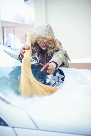 Happy girl cleaning her car from the snow in the city. By the house broom.の写真素材