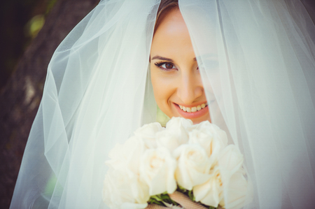 tender happy bride in veil, happy woman in wedding dress with bouquet in hands, white veil covers face, gentle and warm.の写真素材