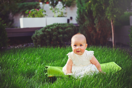 half-year-old child sitting on the grass in the yard, dressed in a white dress rejoices, 6 months. Concept education of children, children's goods.の写真素材