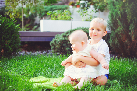 happy little brother playing hugs his sister baby sitting on grass in a green garden, concept of love and parenting.の写真素材