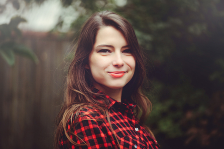 Smiling young brunette woman posing in the courtyard of her residence in a red checkered shirt is...