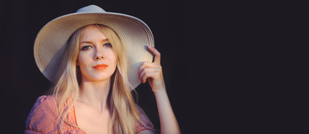 Beautiful young woman in pink dotted summer dress and sun hat points to copy space, looking at...