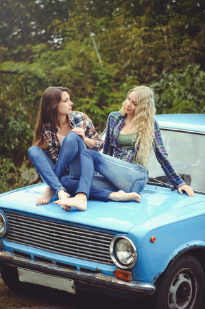 cheerful Attractive two young blonde girl and brunette posing on the hood of an old rusty car, dressed in jeans and shirts on a nature background.の写真素材