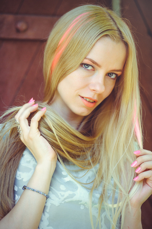 Attractive modest young blond woman playing with hair on red wooden background her hair painted in pink striped striped, in blue dress shy and inquisitive.の写真素材