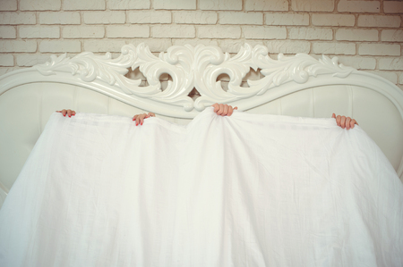 A funny married couple, lying in bed and hiding under a white blanket, only arms are visible. fun in the bedroom. Concept of love and happiness - Imageの写真素材
