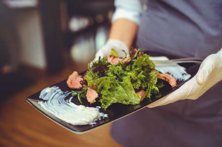 the hands of a waiter in a restaurant with a meat dish with vegetables and herbs.の写真素材