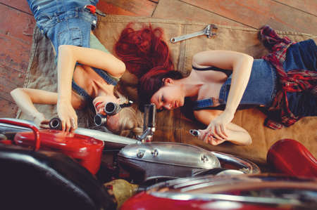 Young beautiful female mechanic working on a custom retro motorcycle. A talented girl in a blue overalls. She uses a ratchet wrench to tighten the nut bolts. Creative Authentic Workshop Garage.の写真素材