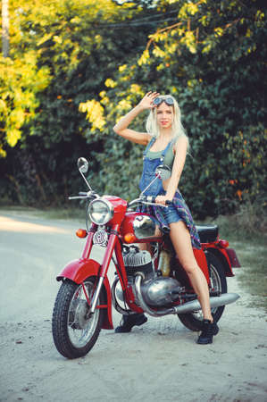 The biker is a beautiful cheerful girl sitting on her motorcycle, a little crazy. Wear short shorts and glasses. holding the steering wheel the wind blows the hair. Looks into the distance at the road.の写真素材