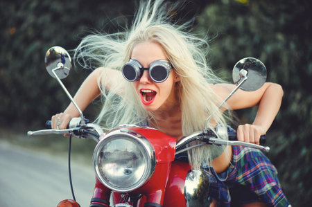 The biker is a beautiful cheerful girl sitting on her motorcycle, a little crazy. Wear short shorts and glasses. holding the steering wheel the wind blows the hair. Looks into the distance at the road.の写真素材