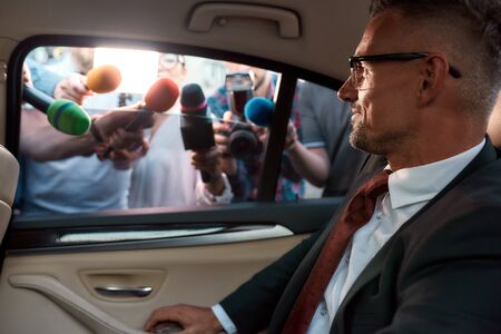 Horizontal portrait of calm man in suit looking at the reporters that attack him holding microphones...