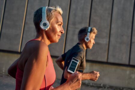 Morning jog. Happy and healthy middle-aged couple in headphones running together through the city...