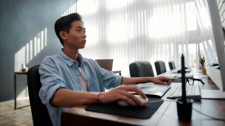 Designer at work. Serious young asian man in casual wear working with computer while sitting at his workplace in the modern office. Creative agencyの写真素材