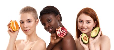 Healthy lifestyle. Three multicultural young and attractive women with glowing skin holding pieces of different fruits and looking at camera while standing against white backgroundの写真素材