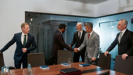 Contract on good conditions. African and asian businessmen in formal wear shaking hands after signing the agreement while standing with their colleagues in the modern officeの写真素材