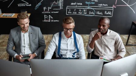 Ideas ahead. Cheerful co-workers traders sitting next to each other in the office and looking at...
