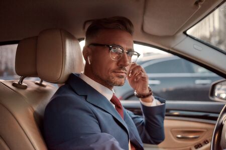 Working from everywhere. Handsome mature man in full suit adjusting headphones while driving a carの写真素材