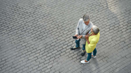 Handsome middle-aged man showing something on smartphone to his wife while standing together outdoors.の写真素材