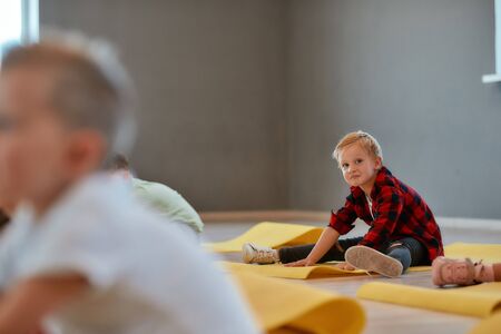 Joyful mood. Portrait of a funny little boy sitting on the yoga mat, stretching and looking at...