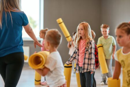 Stretching exercises. Group of cute children in casual clothes holding yoga mats while standing with their female teacher in the dance studioの写真素材