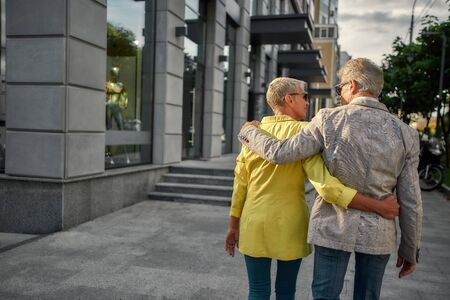 Back view of stylish middle-aged couple in suit jackets are hugging while walking together through the city streetの写真素材