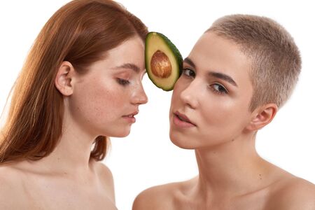 Avocado for beauty care. Young red-haired and blonde women holding avocado between their heads while...