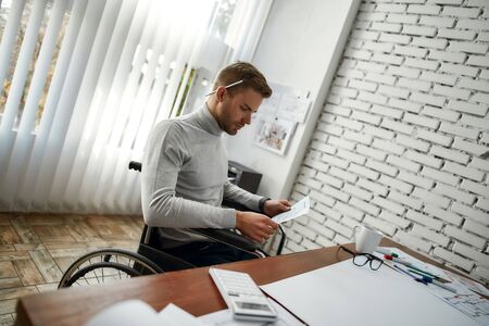 Young and handsome male engineer or architect in a wheelchair looking at blueprint while sitting at his working place in the modern officeの写真素材