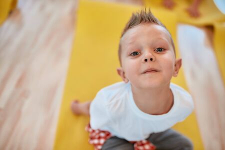 So cute. Portrait of a happy little boy in white t-shirt sitting on the yoga mat and smiling at...