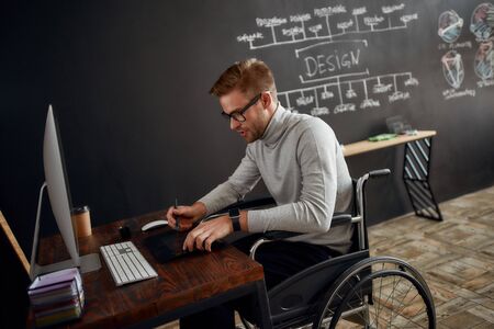 Busy designer at work. Side view of young and handsome male office worker in a wheelchair using stylus pen and digital tablet while sitting at his workplace in design studioの写真素材