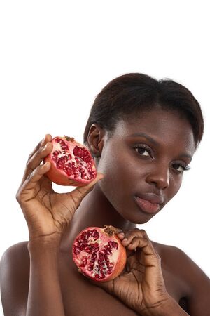 Healthy and tasty. Portrait of beautiful young african woman holding two pieces of fresh juicy pomegranate fruit and looking at camera while standing against white backgroundの写真素材