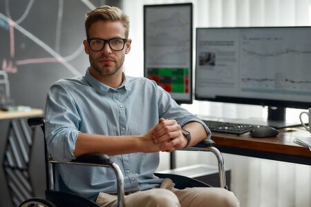 Successful businessman. Portrait of young and confident male trader in a wheelchair looking at camera while working at his workplace in modern office. Disability and handicap conceptの写真素材