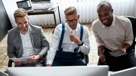 Financial intelligence pays off. Cheerful co-workers traders sitting next to each other in the office and looking at screen of computer while comparing data analyses.の写真素材