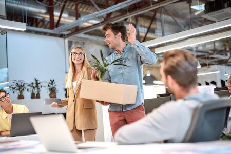 Welcome to our team. Young handsome man in casual wear holding box with things and waving to his new coworkers while standing in the modern officeの写真素材