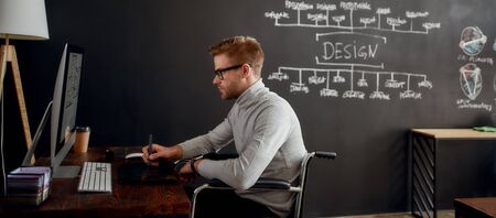 Professional web designer. Side view of young male designer in a wheelchair using stylus pen and digital tablet while sitting at his workplace in creative officeの写真素材
