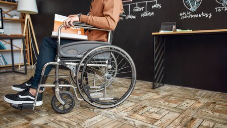 Cropped photo of male office worker in a wheelchair reading a book while sitting near blackboard...