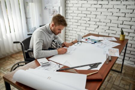 Working day. Young and handsome male engineer or architect in a wheelchair working with blueprint...