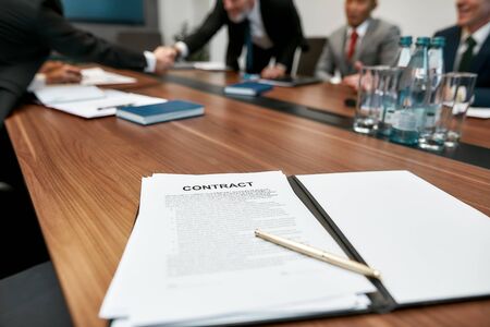 Business agreement. Close up of contract document and pen lying on the office table. Business people in classic wear shaking hands while working together in the modern officeの写真素材